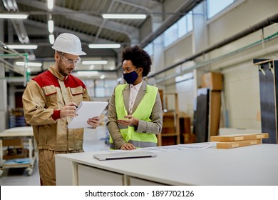 Happy Black Female Engineer Wearing Face Mask While Communicating With Factory Worker At Woodworking Industrial Facility. 