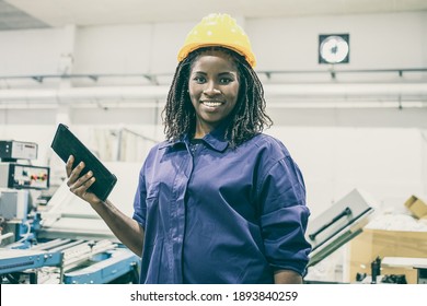 Happy Black Female Factory Worker In Hardhat And Overall, Standing At Machine, Holding Tablet, Looking At Camera And Smiling. Front View, Medium Shot. Women In Industry Concept