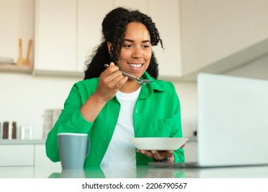 Happy Black Female Freelancer Using Laptop Eating Having Breakfast Sitting At Table In Kitchen At Home. Distance Work And Freelance Career Lifestyle. Selective Focus