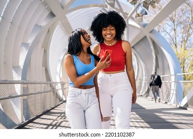 Happy Black Female Friends Laughing And Using Mobile Phone In The City. One Of Them Has Cool Afro Hair.