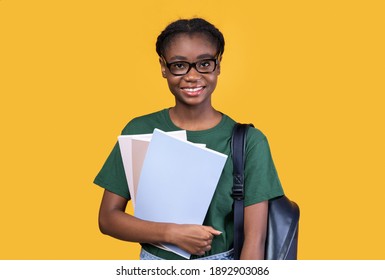 Happy Black Female Student Posing With Books And Notebooks Standing On Yellow Studio Background, Smiling To Camera. College And University Education, Students Lifestyle Concept