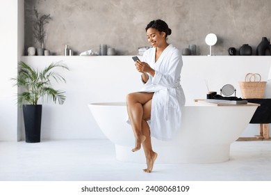 Happy Black Female Using Smartphone While Sitting On Bathtub In Bathroom, Young African American Woman Enjoying Online Communication Or Internet Shopping While Making Daily Beauty Routine At Home - Powered by Shutterstock