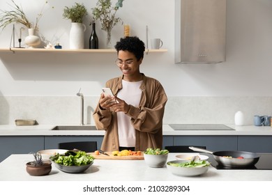 Happy Black food blogger girl taking picture of salad ingredients on mobile phones, recording video, shooting cooking process, reading recipe on Internet, chatting online in home kitchen