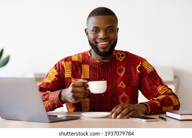 Happy Black Freelancer Guy In African Shirt Using Laptop And Drinking Coffee