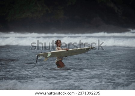Happy black girl holding her surfboard and smiling while standing in the ocean with waves breaking in the background in Sao Tome and Principe, Africa