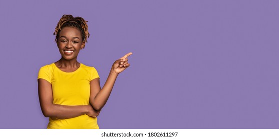 Happy black girl pointing at copy space over purple studio background, panorama, space for advertisement