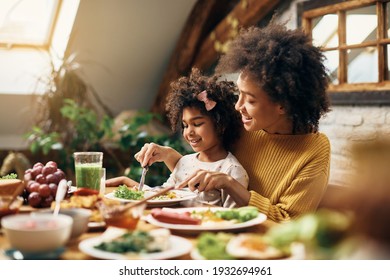 Happy black girl sitting on mother's lap while having a meal at dining table. 