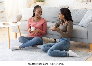 Happy black girlfriends enjoying weekend together, drinking coffee, eating sweets and talking, sitting on floor at home, sharing plans and rumors, copy space, living room interior