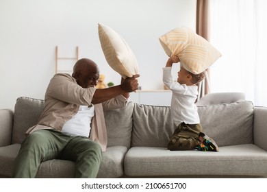 Happy Black Grandfather And Grandchild Boy Having Pillow Fight For Fun Spending Weekend At Home. Grandpa And Little Grandson Playing Together Fighting With Pillows Indoor