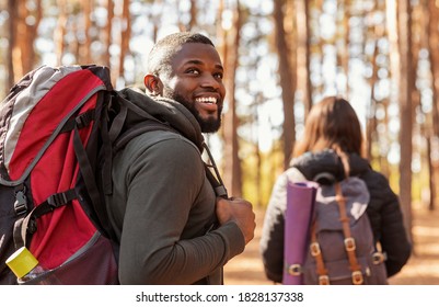 Happy black guy with backpack looking aside, hiking with girlfriend at forest