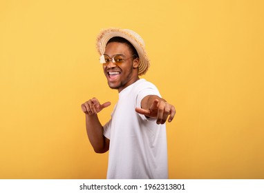 Happy Black Guy In Casual Summer Outfit, Straw Hat And Sunglasses Pointing At You On Yellow Studio Background. Positive African American Man Enjoying Summertime, Having Fun