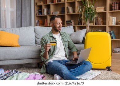 Happy Black Guy With Credit Card Booking Vacation Or Making Hotel Reservation Online, Using Laptop, Sitting Near Suitcase At Home. Man Planning Abroad Trip