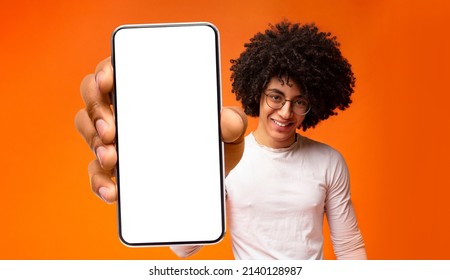 Happy black guy demonstrating smartphone with big blank white screen, showing free copy space for your ad, app or website design, standing over orange background, mockup