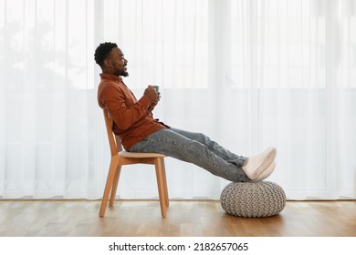 Happy Black Guy Drinking Coffee Holding Mug Sitting In Chair Relaxing At Home. Man Resting Enjoying Hot Drink Near Window In Modern Living Room Indoor. Weekend Lifestyle Concept. Side View