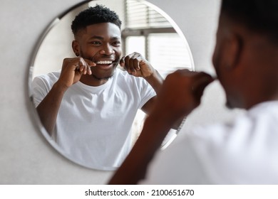 Happy Black Guy Flossing Teeth With Tooth Floss Doing Daily Oral Hygiene Routine, Looking At Mirror In The Morning Standing In Modern Bathroom Indoors. Dental Health And Toothcare Concept