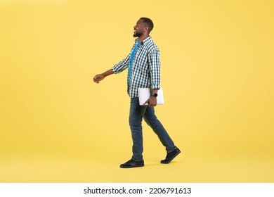 Happy Black Guy Holding Laptop Walking Looking Aside Over Yellow Studio Background. Internet Business And Freelance Career Concept. Side View, Full Length Shot