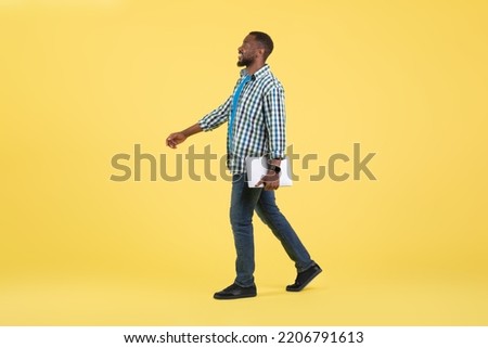 Happy Black Guy Holding Laptop Walking Looking Aside Over Yellow Studio Background. Internet Business And Freelance Career Concept. Side View, Full Length Shot