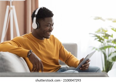 Happy Black Guy Sitting On Couch, Watching Movies On Digital Tablet At Home, Copy Space. Smiling African American Young Man Reclining On Sofa, Using Modern Pad And Wireless Headphones, Side View