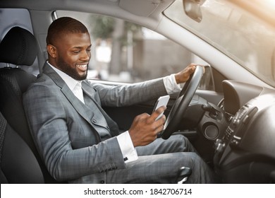 Happy Black Guy In Suit Using Smartphone While Driving Car. Positive African American Businessman Chatting With Friends Or Clients On Mobile Phone While Going To Office By Car, Copy Space