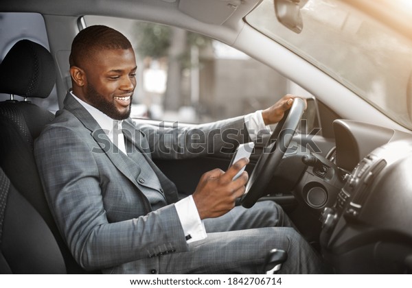 Happy black guy in suit using smartphone while\
driving car. Positive african american businessman chatting with\
friends or clients on mobile phone while going to office by car,\
copy space