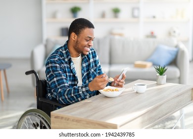 Happy Black Guy In Wheelchair Eating Breakfast And Browsing Web On Smartphone At Home, Copy Space