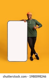 Happy Black Hipster Guy Leaning At Big Smartphone With Blank White Screen And Gesturing Thumb Up, Cheerful African Man Recommending New App Or Website, Standing On Yellow Background, Mockup Image