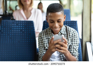Happy Black Kid Using Cell Playing Mobile Games Online On Smartphone Connected To Public Wifi Sitting On Seat In City Bus. Smiling Teen Passenger Looking At Device Gadget Screen, Texting Sms