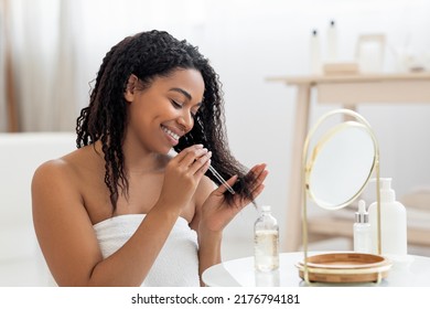 Happy Black Lady Applying Moisturising Oil On Damaged Hair Ends After Shower, Smiling African American Woman Sitting Near Mirror In Bathroom Interior, Making Haircare Routine At Home, Closeup