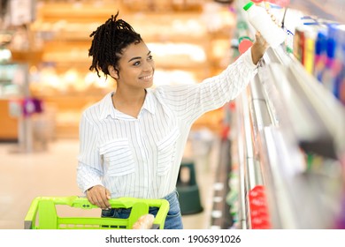 Happy Black Lady Buying Food Walking With Shop Cart In Modern Supermarket, Taking Dairy Product From Shelf In Grocery Shop. Lady Choosing Groceries Buying Supplies. Selective Focus