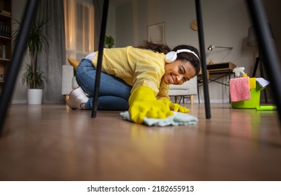 Happy Black Lady Dusting And Mopping Floor With Rag Under Table Cleaning Modern Living Room At Home, Wearing Rubber Gloves. Female Doing House Chores. Selective Focus