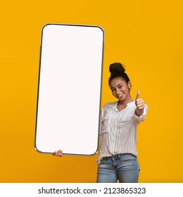 Happy Black Lady Holding Big Smartphone With Blank Screen And Showing Thumb Up, Recommending Educational Mobile Application Or Website, Orange Background, Mockup