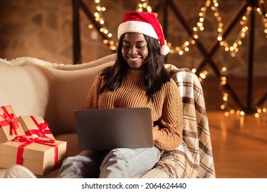 Happy Black Lady In Santa Hat Using Laptop, Ordering Xmas Gifts On Web, Shopping For Christmas Presents Online From Home. African American Woman Sitting On Couch With Computer On Winter Holidays