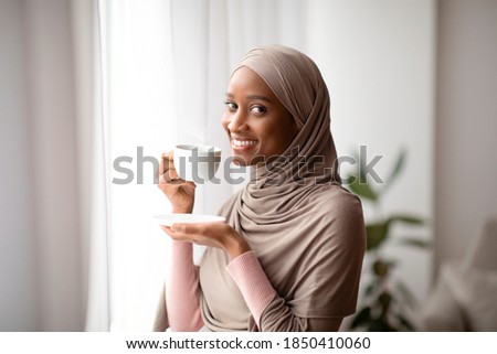 Happy black lady in traditional hijab drinking fresh coffee near window at home, enjoying relaxing morning. Portrait of African American woman in headscarf having hot beverage indoors
