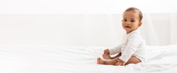 Joyeuse Petite Bébé Noire Assis Sur Un Lit Regardant La Caméra Et Souriant Dans La Chambre À La Maison. Bannière Publicitaire Avec Un Joli Enfant Posant À L'Intérieur. Panorama Avec Espace Pour Copier Votre Texte