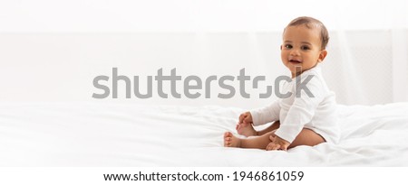 Happy Black Little Baby Girl Sitting On Bed Looking At Camera And Smiling In Bedroom At Home. Advertisement Banner With Cute Child Posing Indoor. Panorama With Copy Space For Your Text