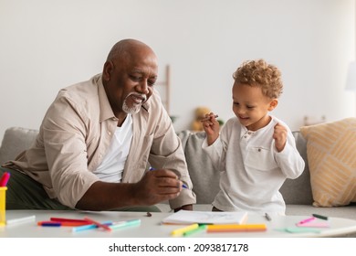 Happy Black Little Boy And His Senior Grandpa Drawing Picture With Felt Pens Sitting On Sofa At Table At Home. Grandfather Spending Time With Grandson Sketching Together On Weekend