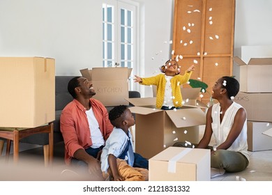 Happy black little girl playing with polystyrene foam chips while standing inside a box. Happy african family enjoying new home. Parents with son and funny daughter having fun during moving house.
