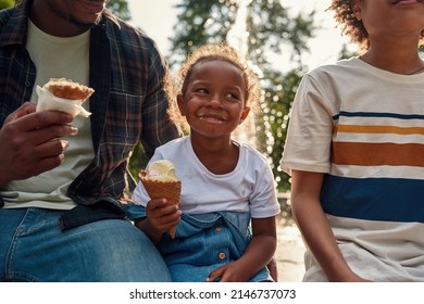 Happy Black Little Girl Resting And Eating Ice Cream Cones With Her Father And Brother Near Fountain In Park. Family Relationship And Enjoying Time Together. Fatherhood And Parenting. Sunny Summer Day