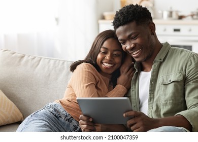 Happy black lovers millennial man and woman sitting on couch, hugging and using digital tablet together, looking at pad screen and smiling, using newest mobile application, home interior, copy space - Powered by Shutterstock