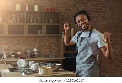 Happy black man in earphones listening to music and dancing while baking in kitchen