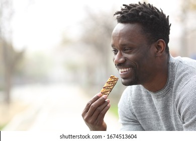 Happy Black Man Eating A Cereal Bar In A Park Looking Away