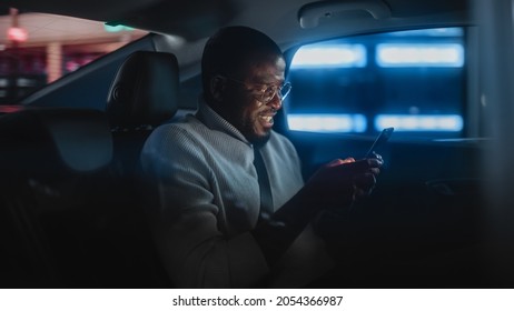 Happy Black Man In Glasses Is Commuting Home In A Backseat Of Taxi At Night. Handsome Male Using Smartphone And Smiling While In A Car In Urban City Street With Working Neon Signs.