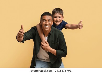 Happy black man have fun posing with cute child preteen boy. Daddy and son piggybacking and showing thumb ups, beige background studio portrait. Fathers Day love family parenthood childhood concept - Powered by Shutterstock