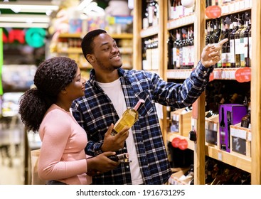 Happy black man and his girlfriend buying wine at liquor department of supermarket. Lovely African American couple shopping for alcoholic drinks at huge mall together