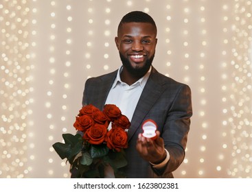 Happy black man holding red roses and box with wedding ring, proposal concept