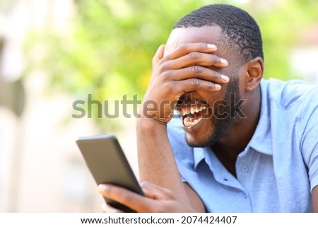 Happy black man laughing loud checking smart phone content in a park