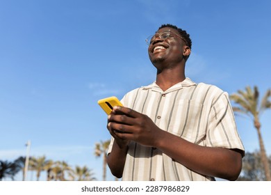 Happy black man laughing outdoors using mobile phone. Blue sky background. Copy space.