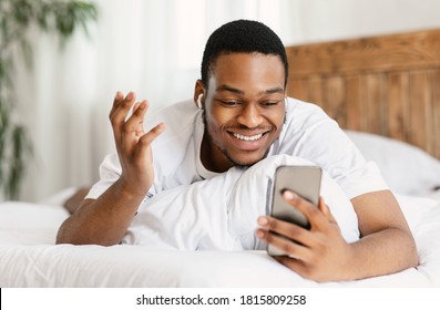 Happy Black Man Making Video Call On Cellphone Using Smartphone And Earbuds Earphones For Distant Communication Lying In Bed In Bedroom At Home On Weekend. Selective Focus - Powered by Shutterstock