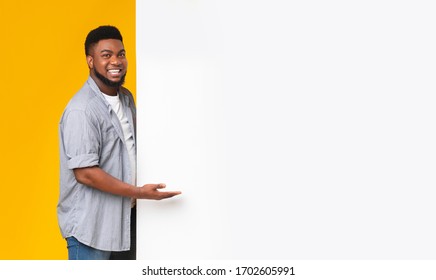 Happy black man pointing at blank white advertisement board with open palm, standing over yellow background