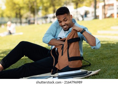 Happy Black Man Searching Something In His Backpack, Sitting On Grass In Park Outdoors. African American Male Student Getting Something Out Of His Bag And Smiling